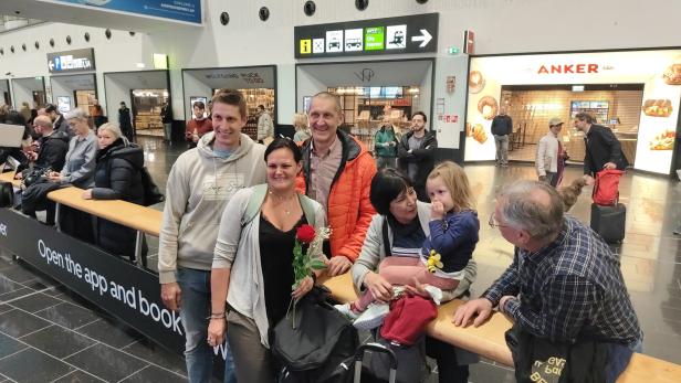 Sechs Menschen, darunter ein kleines Kind, stehen und sitzen lächelnd zusammen in einer belebten Bahnhofshalle.