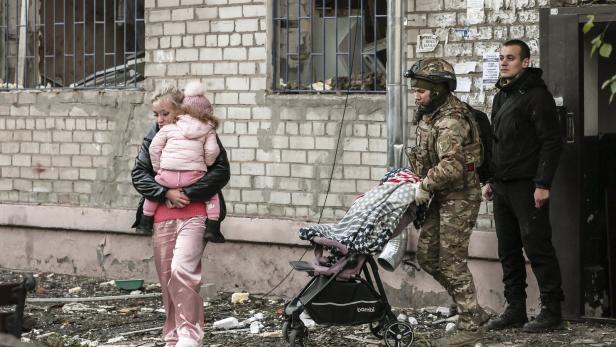 Eine Frau mit Kind, ein Soldat mit Kinderwagen und ein Mann stehen vor einem beschädigten Gebäude mit zerbrochenen Fenstern.
