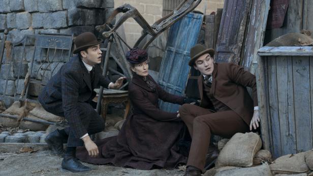 Drei Personen in historischer Kleidung ducken sich hinter Barrikaden aus Holz und Sandsäcken und wirken angespannt.