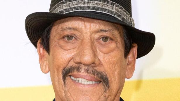Danny Trejo mit Hut und Jeansjacke mit Aufnähern lächelt.