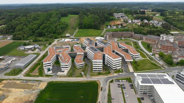 Mehrere moderne Gebäude mit roten Dächern und großen Fenstern liegen inmitten grüner Landschaft und Waldflächen.