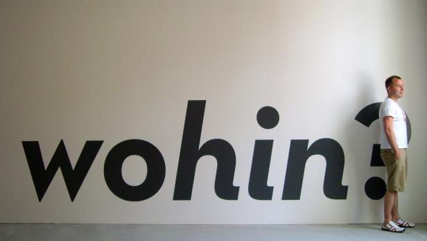 Ein Mann steht vor einer weißen Wand mit dem großen schwarzen Schriftzug „wohin?“ und blickt nach rechts.