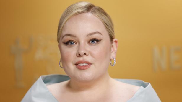 Nicola Coughlan mit blonden Haaren und hellblauen Augen lächelt vor einem gelben Hintergrund.