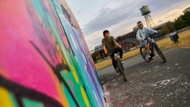 zwei Personen fahren mit dem Fahrrad neben einer Wand, die mit Graffiti bemalt ist