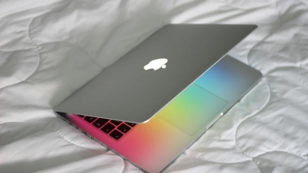 Ein teilweise geöffnetes MacBook mit sichtbarem Apple-Logo liegt auf einer weißen Decke, das Display wirft bunte Lichtreflexe auf die Tastatur.