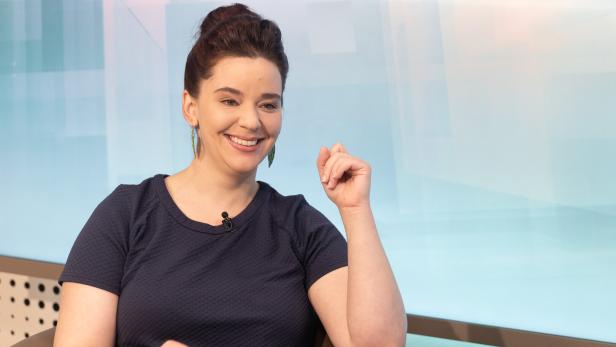 Eine Frau mit dunklen Haaren sitzt lächelnd auf einem Sessel und trägt ein dunkelblaues Oberteil.