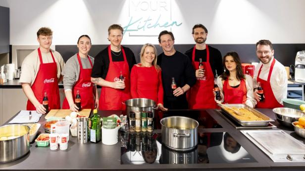 Acht Personen mit roten Coca-Cola-Schürzen stehen lächelnd in einer modernen Küche.