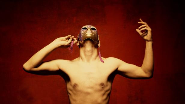 Oberkörper einer Person mit ausgebreiteten Armen und gold-violetter Maske vor rotem Hintergrund.