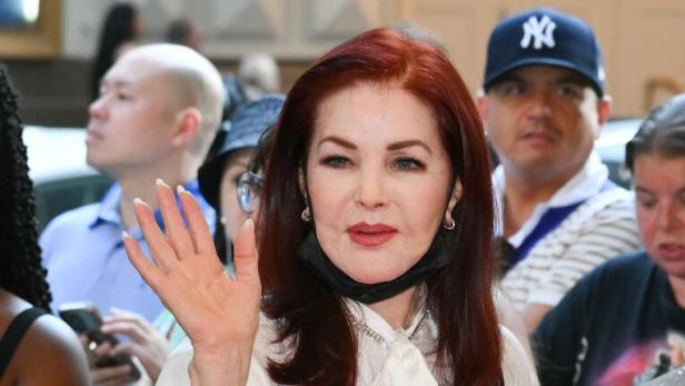 Priscilla Presley mit rotem Haar, weißer Bluse und schwarzem Rock winkt in die Menge.