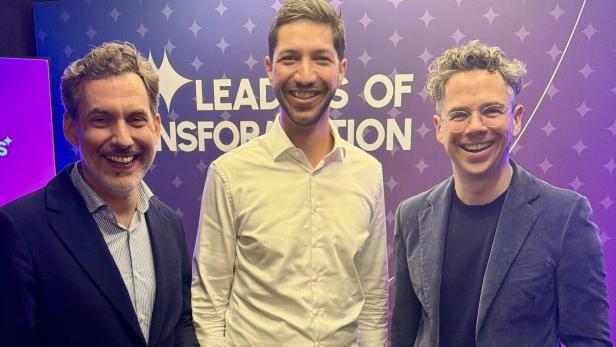 Drei Männer stehen lächelnd vor einer lila Wand mit der Aufschrift „Leaders of Transformation“.