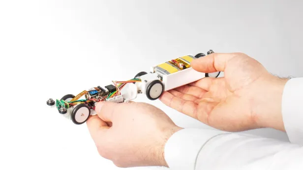 2 Hände halten ein kleines, selbstgebautes Roboterfahrzeug mit sichtbaren elektronischen Bauteilen und Kabeln.