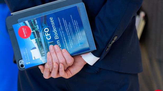 Man sieht eine Person von hinten, ihre Hände sind nah rangezoomt. Sie hält ein Notizbuch und einen Flyer in der Hand auf der man die Ankündigung des CEO/CFO-Forums sieht.