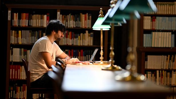 Ein Mann sitzt an einem langen Tisch in einer Bibliothek und arbeitet an einem Laptop unter einer grünen Lampe.