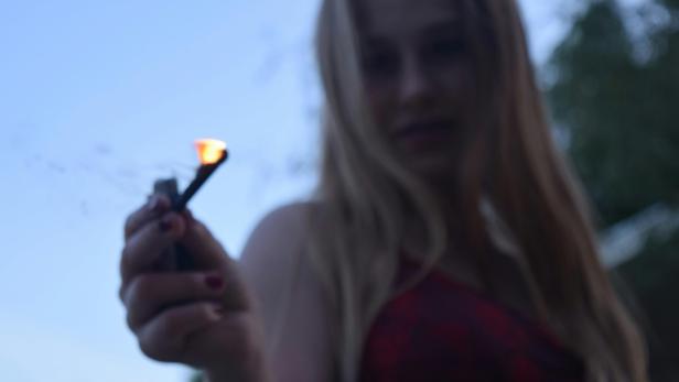 Ein Mädchen hält ein brennendes Streichholz in der Hand, offenbar im Freien, mit unscharfem Hintergrund.