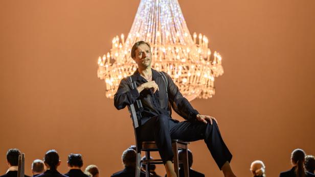 Ein Mann sitzt auf einem Stuhl vor einem großen Kronleuchter, im Hintergrund mehrere Personen mit dem Rücken zur Kamera.
