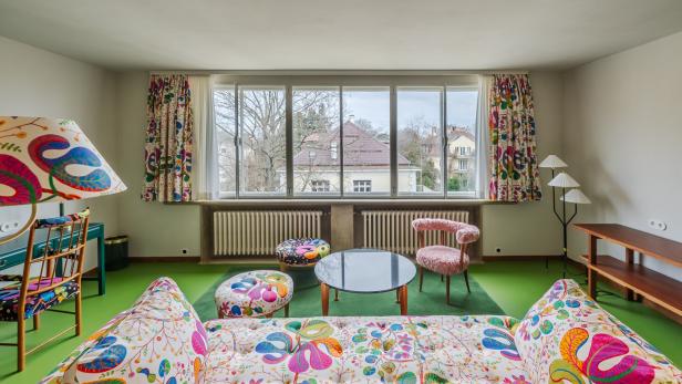 Wohnzimmer mit großen Fenstern, bunten Möbeln und Vorhängen, grünem Boden und Blick auf Häuser und Bäume draußen.