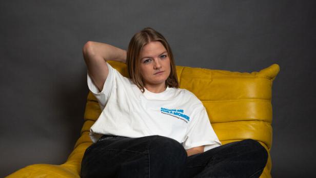 Junge Frau sitzt entspannt auf einem gelben Sessel, trägt weißes T-Shirt, schwarze Hose und weiße Sneaker.