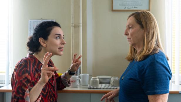 Zwei Frauen stehen sich in einer Küche gegenüber, eine spricht gestikulierend mit erhobenen Fingern, die andere hört aufmerksam zu.