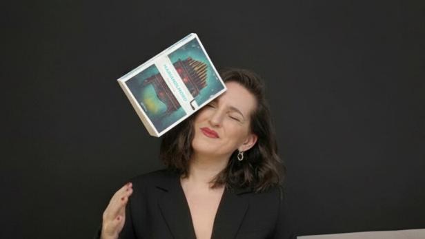 Eine Frau in schwarzer Kleidung sitzt auf einem Sofa, während ihr ein Buch ins Gesicht fällt.