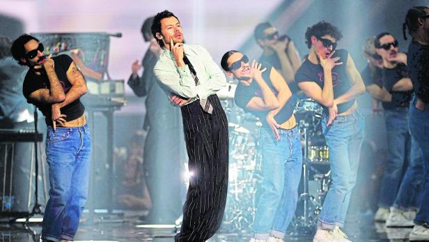 Mehrere Tänzer und ein Sänger - Harry Styles - in auffälliger Pose auf einer Bühne mit Lichteffekten und Instrumenten im Hintergrund.