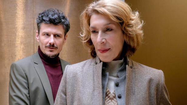 Eine Frau mit blondem Haar steht im Vordergrund, ein Mann mit dunklem Haar blickt sie ernst von hinten an.