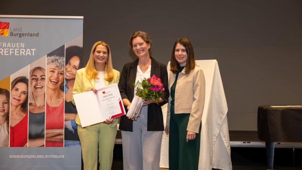Drei Frauen stehen lächelnd zusammen, eine hält eine Urkunde, eine andere einen Blumenstrauß.