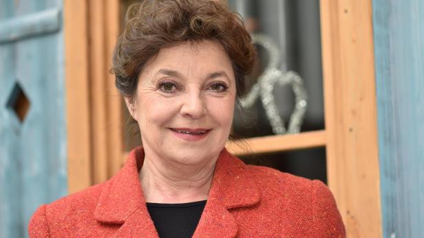 Eine lächelnde Frau mit braunem Haar trägt ein rotes Jackett vor einem Fenster.