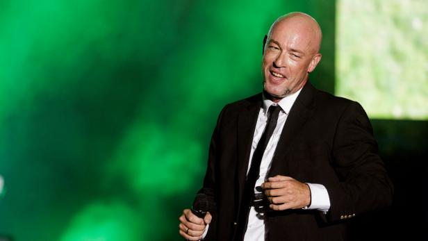 Der Graf von Unheilig steht im schwarzen Anzug mit Mikrofon auf einer Bühne vor grünem Hintergrund.