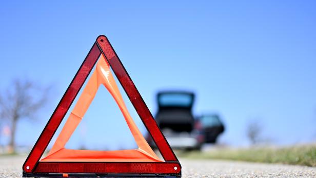 Ein rotes Warndreieck steht auf einer Straße, im Hintergrund ist ein Auto mit offenem Kofferraum zu sehen.
