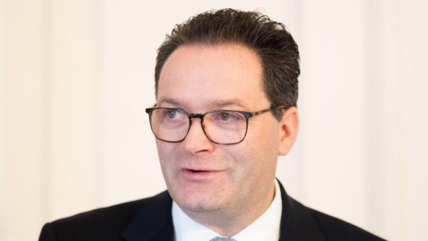 Mann mit dunklem Anzug, weißem Hemd, gemusterter Krawatte und Brille vor hellem Hintergrund.