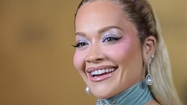 Rita Ora mit glänzendem Make-up, auffälligem Schmuck und hellblauem Samtband um den Hals lächelt.
