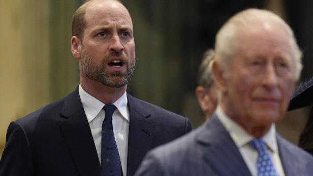 Prinz William im Hintergrund, König Charles im Vordergrund. Beide tragen Anzüge und schauen ernst.