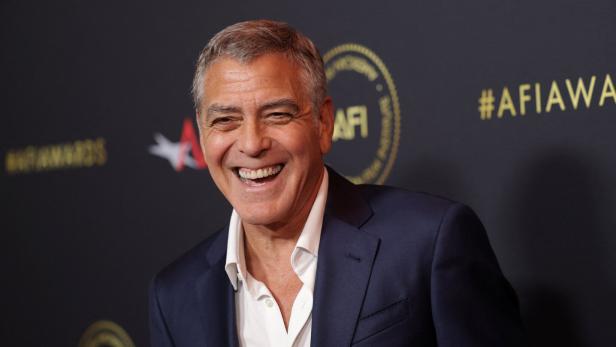 George Clooney im dunklen Anzug und weißem Hemd lächelt vor einer schwarzen Wand mit goldener Schrift.