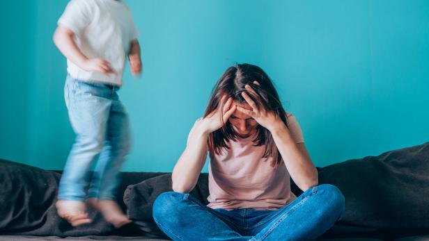 Eine Frau greift sich an den Kopf, während ein Kind auf dem Sofa hüpft.