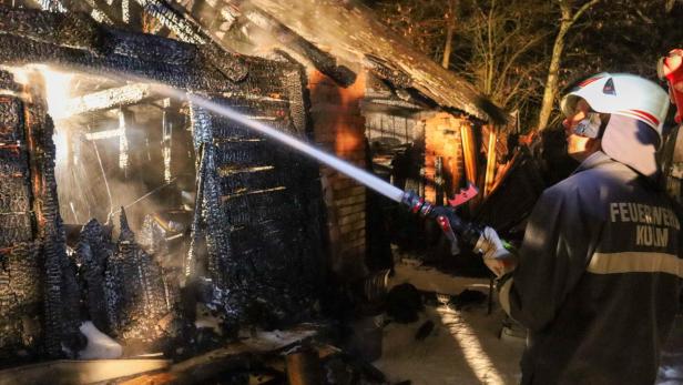 Ein Feuerwehrmann löscht mit einem Wasserschlauch ein stark verbranntes, teilweise zerstörtes Gebäude.