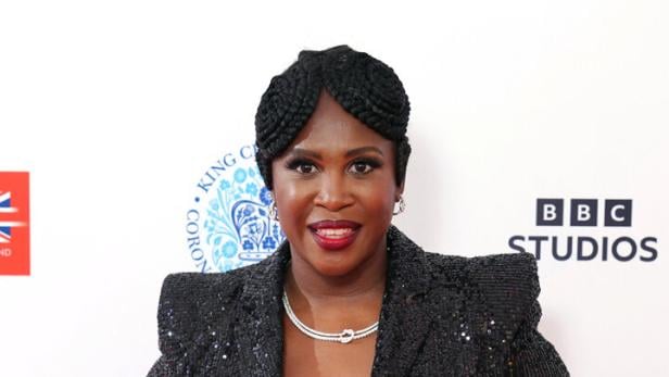 Motsi Mabuse mit geflochtener Frisur trägt einen schwarzen, glitzernden Anzug und Schmuck, posiert vor einer Sponsorenwand.