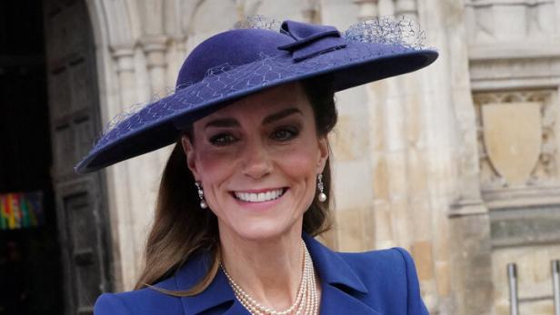 Kate trägt einen blauen Hut und Blazer und hält einen Blumenstrauß.