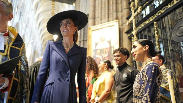 Eine elegant gekleidete Frau mit großem Hut steht in einer prunkvollen Kirche neben weiteren festlich gekleideten Personen.