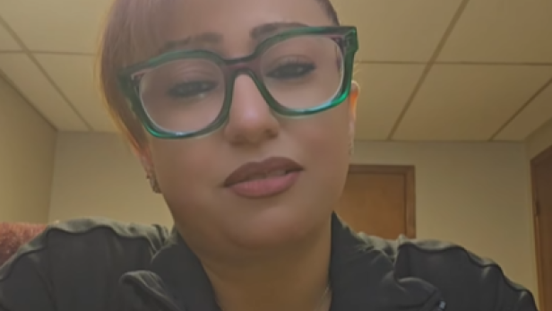 Frau mit Brille und kurzem Haar sitzt in einem Raum mit Holzdecke und geschlossenen Türen.