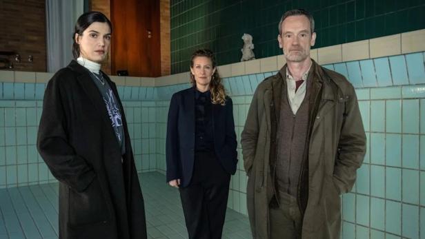 Drei Personen stehen in einem leeren, gekachelten Schwimmbecken und blicken ernst in die Kamera.