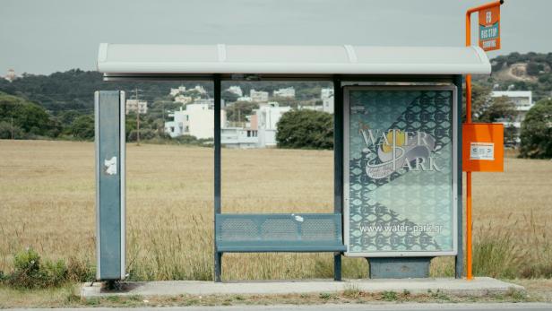 Leere Bushaltestelle an einer Landstraße mit Feldern und Gebäuden im Hintergrund.