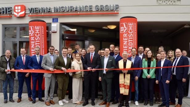 Eine Gruppe von Menschen steht vor einem Versicherungsbüro und durchschneidet gemeinsam ein rotes Band zur Eröffnung.