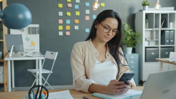 Eine Frau sitzt an einem Schreibtisch in einem modernen Büro und liest konzentriert auf ihrem Smartphone, im Hintergrund bunte Notizzettel an einer Wand.