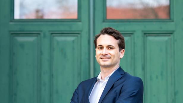 Ein Mann im blauen Anzug steht mit verschränkten Armen vor einer großen grünen Holztür und lächelt.