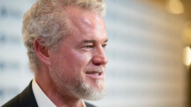 Eric Dane mit grauem Haar und Bart, gekleidet in einem dunklen Anzug, blickt nach rechts. 