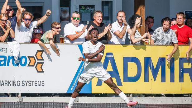 Ein Fußballspieler (Alieu Conateh) jubelt vor begeisterten Fans, die am Spielfeldrand klatschen und jubeln.