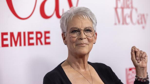 Jamie Lee Curtis lächelt mit Brille vor einem roten Schriftzug auf weißem Grund.