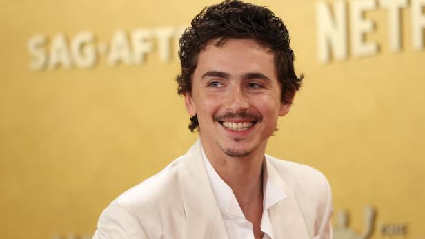 Ein lächelnder Timothée Chalamet mit dunklen Locken und weißem Anzug vor goldenem Hintergrund.