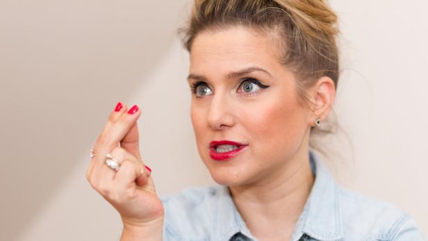 Jeanette Biedermann mit rotem Lippenstift gestikuliert mit den Fingern vor einem hellen Hintergrund.