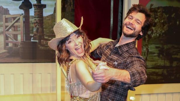 Ein lachendes Paar tanzt, die Frau trägt einen glitzernden Fransenanzug und Cowboyhut, der Mann ein kariertes Hemd.